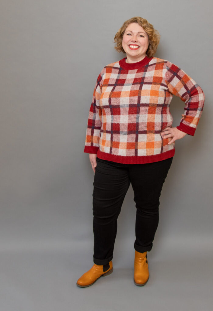 Sarah smiling, standing with her hand on her hip, wearing a red plaid sweater, black pants, and yellow boots. 