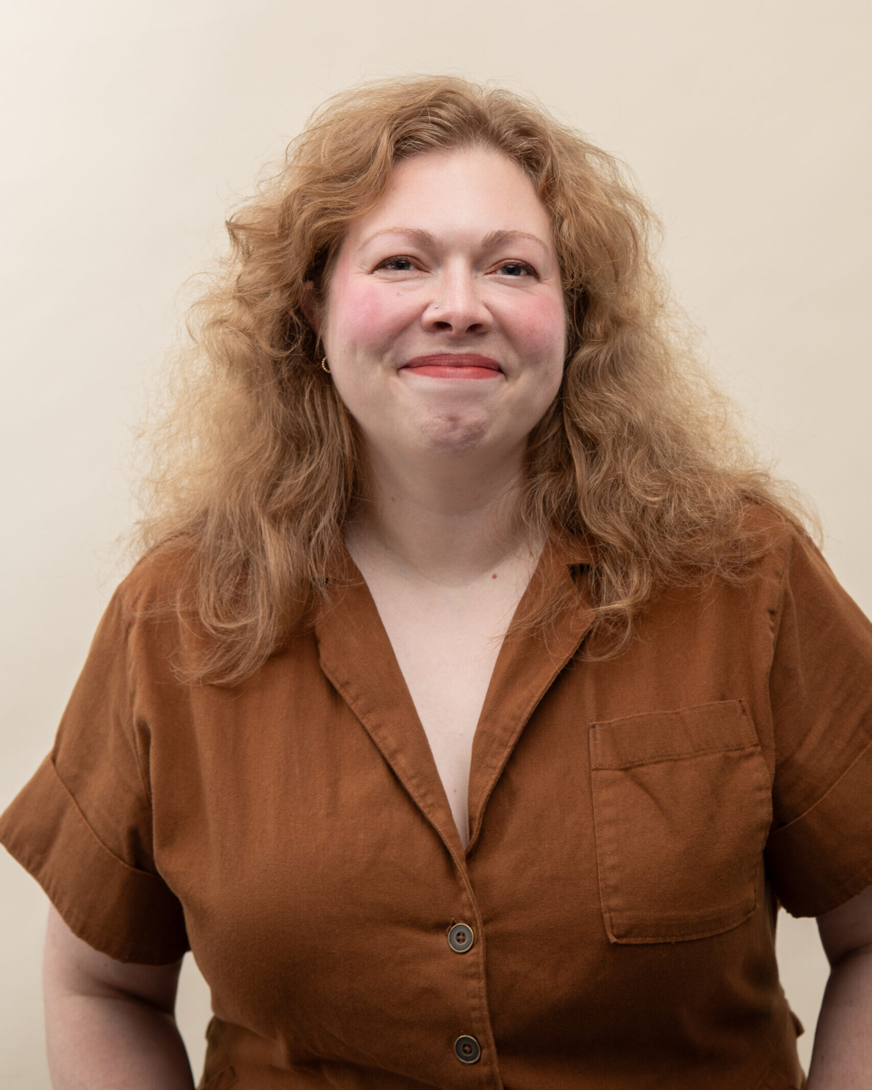 A white woman with wavy blond hair is facing the camera and smiling. She is wearing a brown shirt.